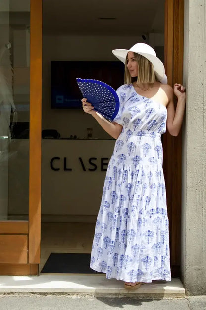modella bionda indossa un abito lungo estivo monospalla con ventaglio blu a pois bianche e cappello di paglia blu